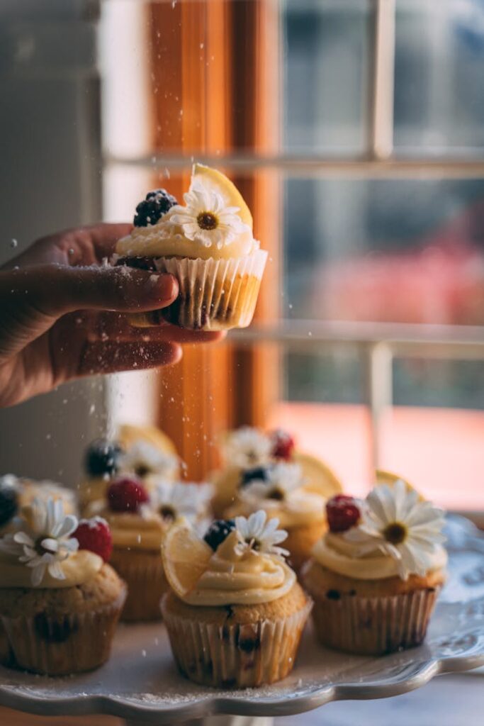 Hand holding a lemon and berry cupcake with flower decoration in a cozy setting.