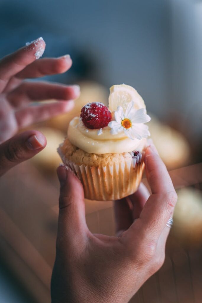 Close-up of a cupcake topped with raspberry, cream, and edible flower held by hands.