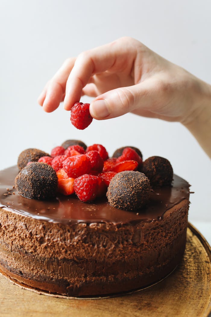 Close-up of a hand decorating a chocolate cake with raspberries and truffles.